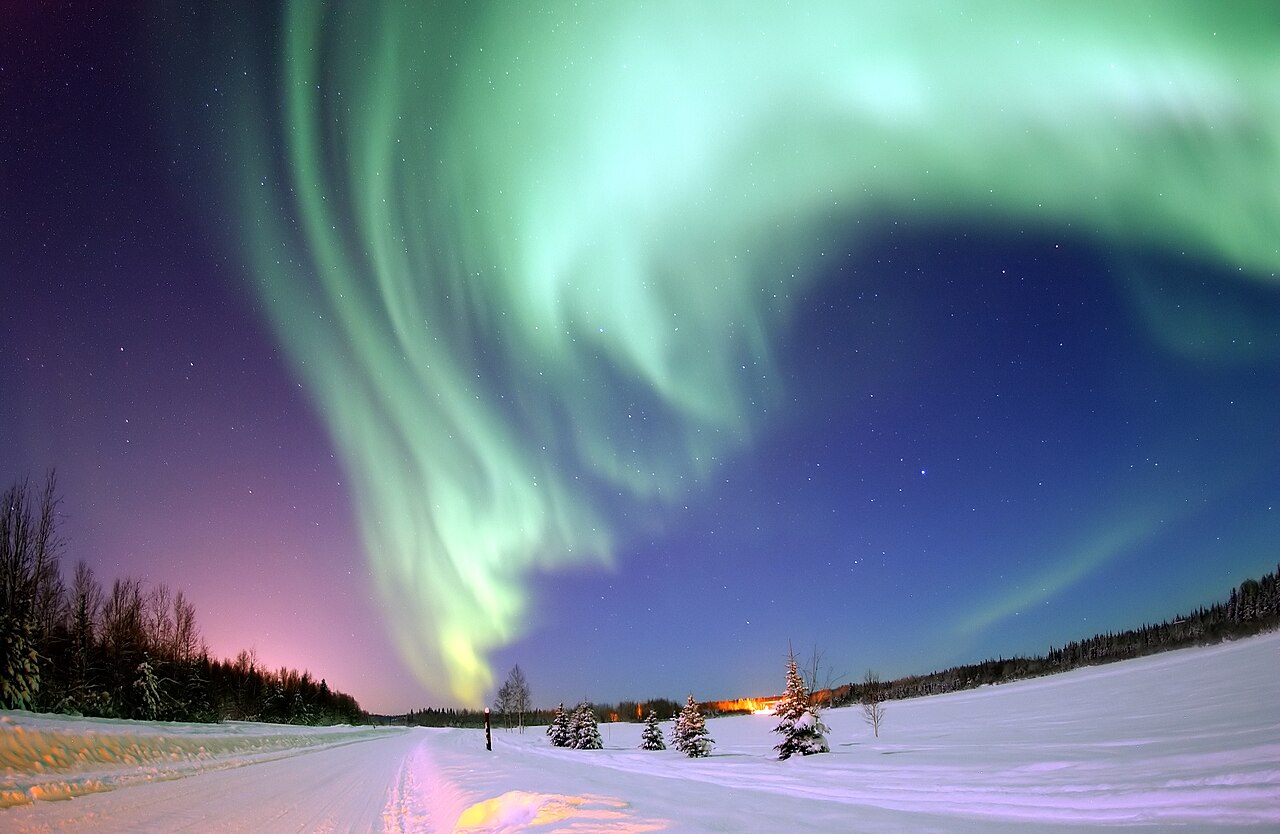 Green aurora borealis curtains arching over a frozen lake and a dark tree line near Fairbanks, Alaska