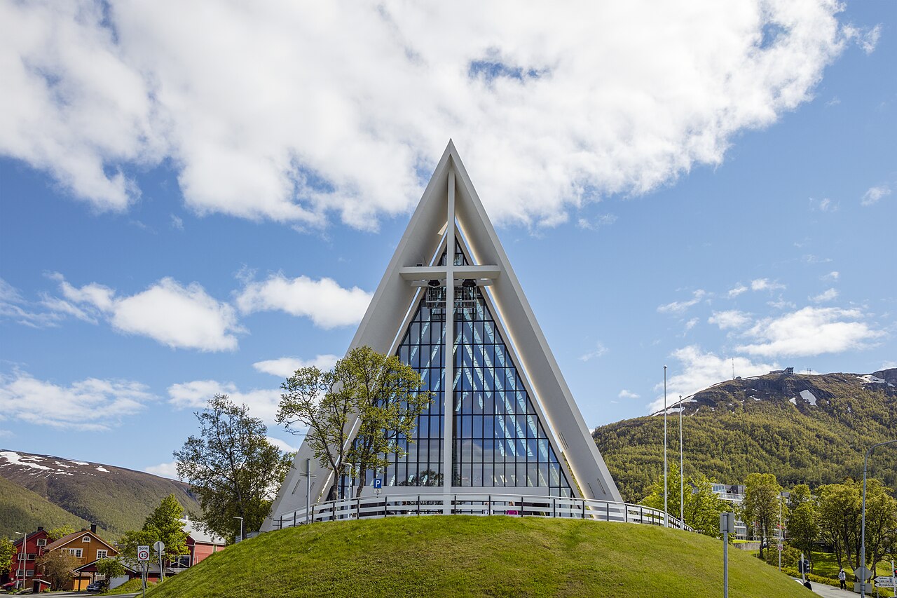 The white triangular Arctic Cathedral with its large glass facade on a green hill under a blue sky